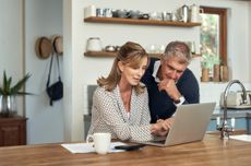 A senior couple sits at the kitchen table in a modern home working on a laptop.